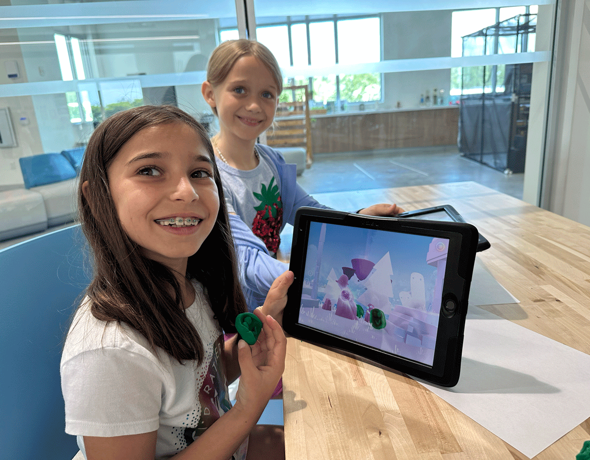 Two girls sit at a table. One holds green clay, while the other displays a tablet showing a digital scene. Both are smiling.