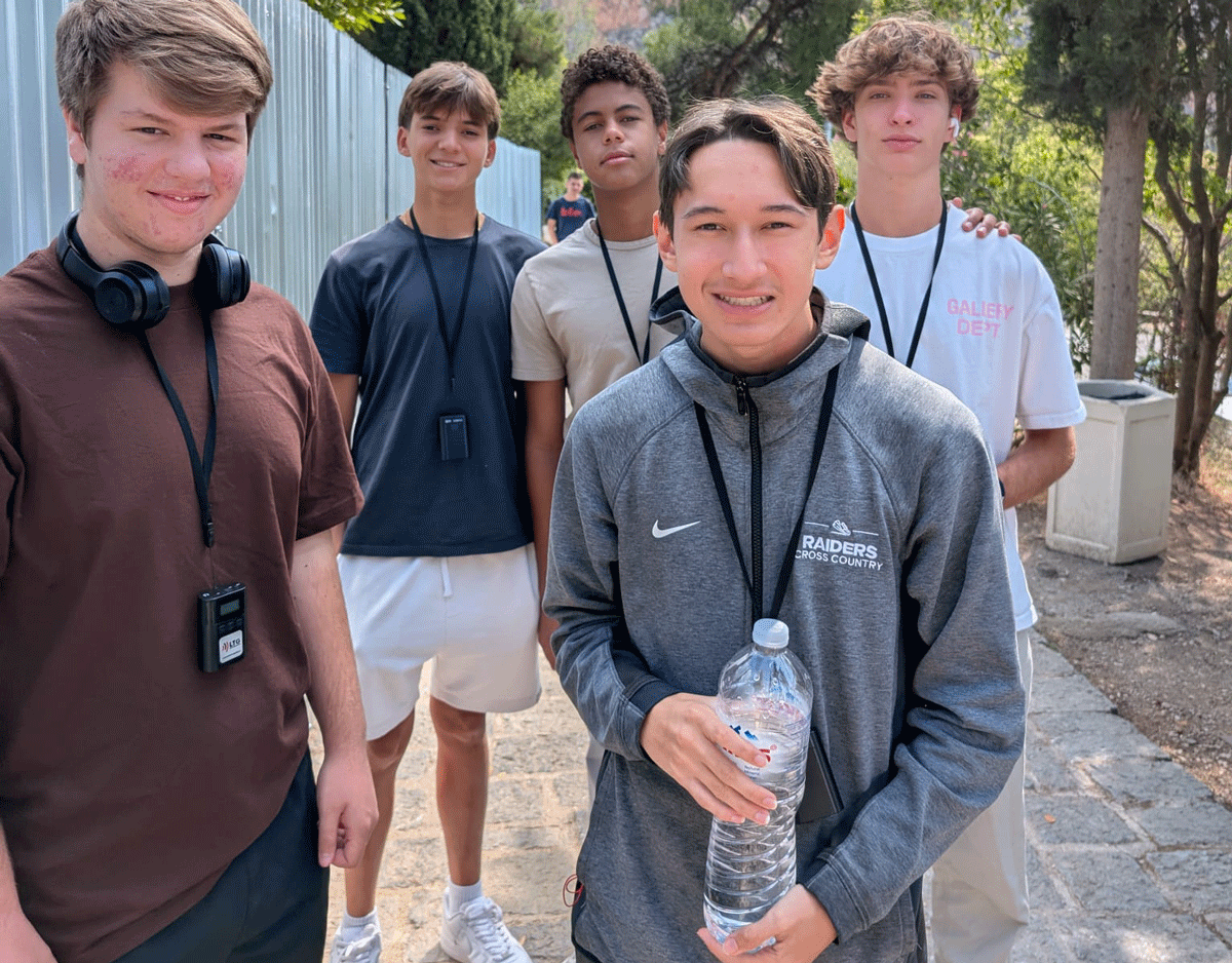 Five teenage boys stand outdoors on a paved path. One holds a water bottle. Trees and a fence line the background. The group appears relaxed and casually dressed.