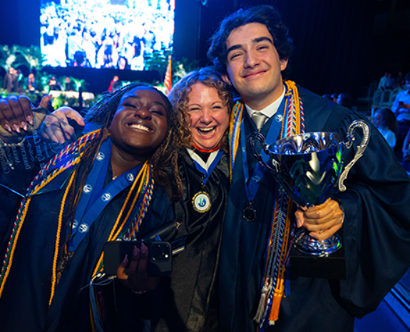 Three graduates in caps and gowns smile and celebrate, holding a trophy and medals, with a crowd and stage in the background.