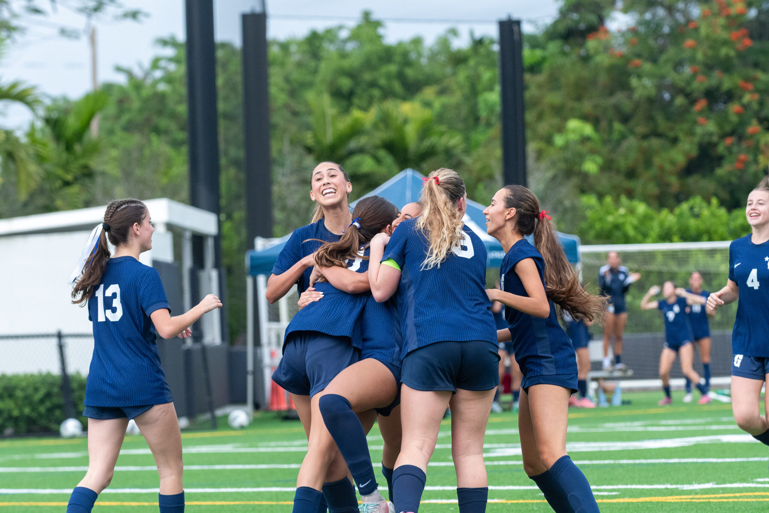 A group of female soccer players in blue uniforms celebrate together on a field, hugging and smiling, with trees and a fence in the background.