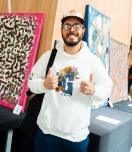 An alumni spotlight: a man in a white graphic hoodie and a baseball cap smiles and gives a double thumbs-up in front of displayed art pieces.