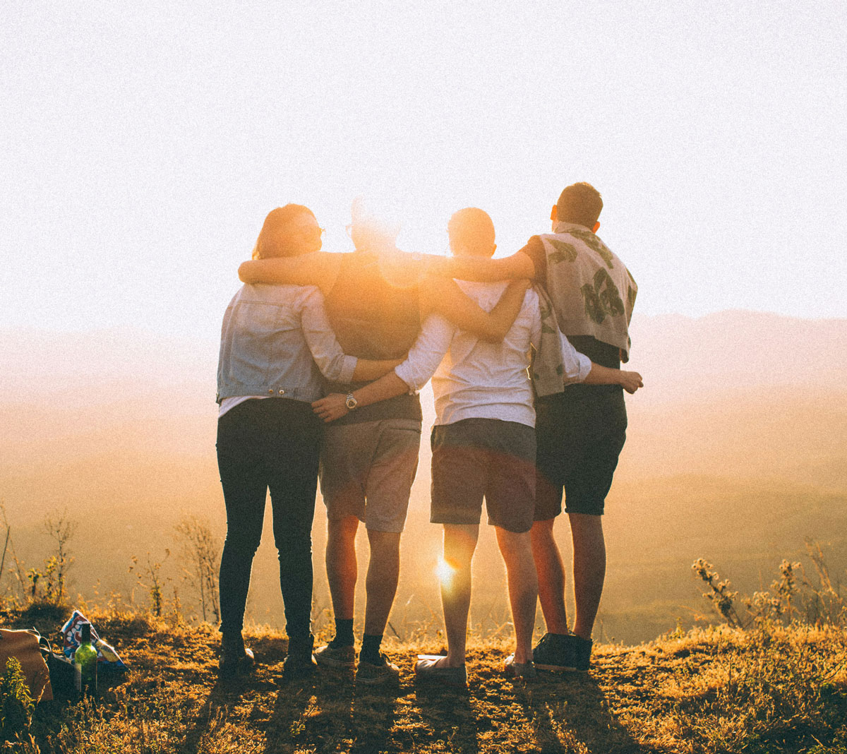 Four people stand together outdoors with arms around each other, facing a sunset or sunrise over a scenic landscape, sharing a moment of connection often found among colleagues pursuing private school careers.