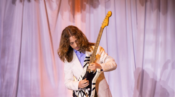 A young person with long hair wearing a white blazer plays a black and white striped electric guitar on stage in front of a curtain.